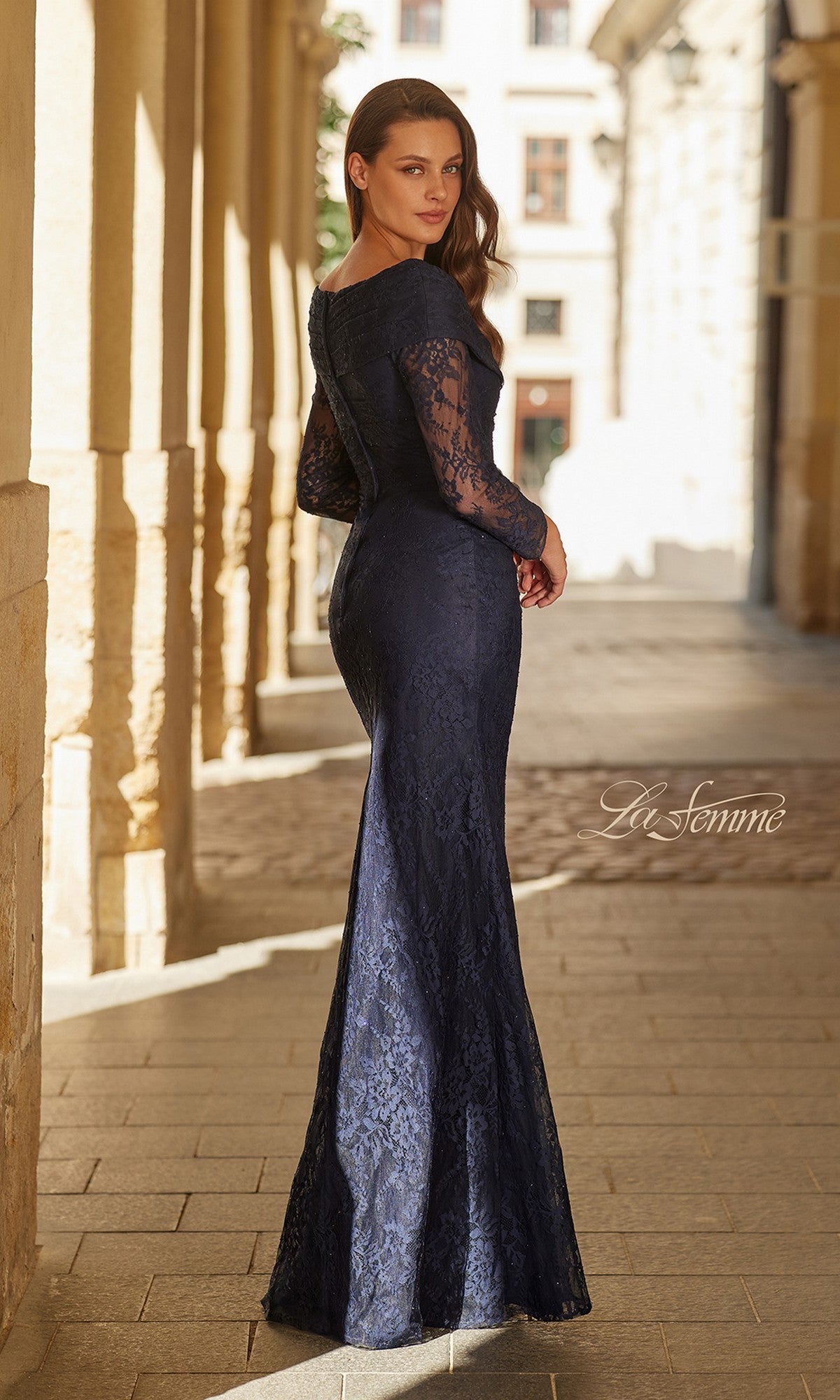 A woman stands in a sunlit outdoor corridor, looking over her shoulder in the fitted, floor-length La Femme 33333 long formal dress with navy lace and long sleeves.